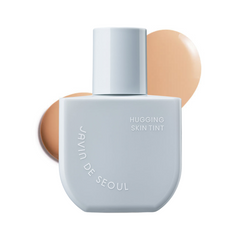 Lysegrå flaske med "Javin de Seoul Hugging Skin Tint" og brune farveprøver i baggrunden.